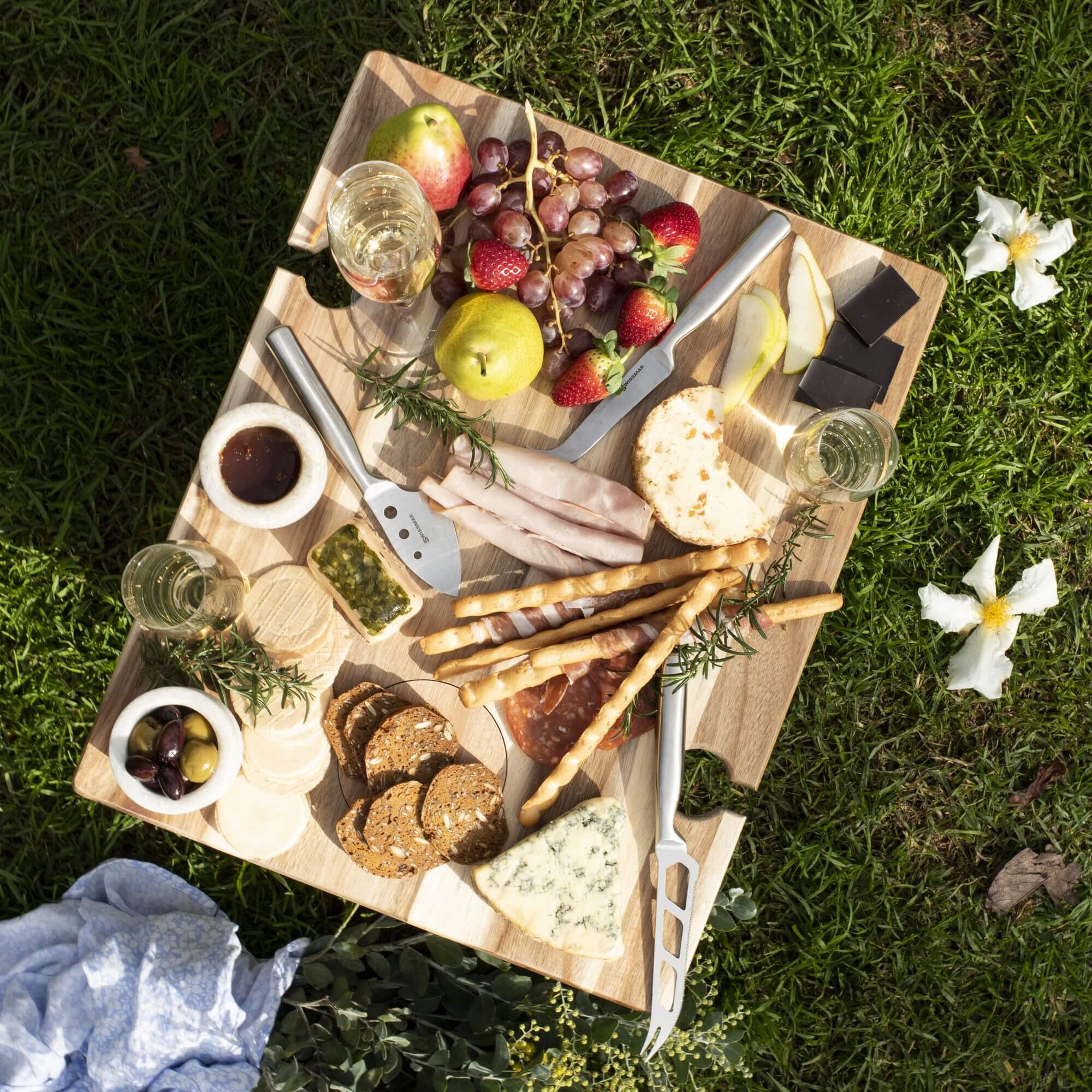 A bamboo serving board with wine glasses and cheese knives